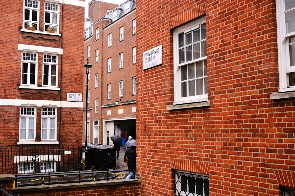 The image shows the exterior of a red brick building on a street corner in Marylebone, Westminster, with visible signage including 'Providence Court W1' and 'Balderton Street W1'. Two movers, dressed