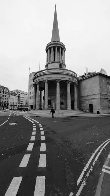 Two cyclists wearing helmets ride along a paved street in Marylebone, outside a large white neoclassical building with tall columns and decorative railings. In the background, a group of pedestrians i
