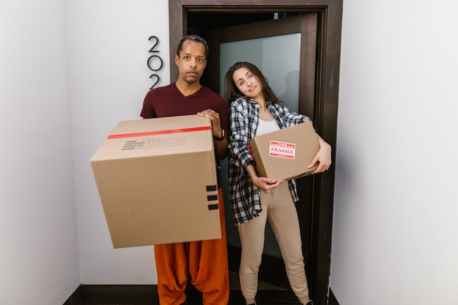 A man and a woman standing inside an entryway near a doorway in an indoor property, each holding a cardboard box used for packing and moving; the man, wearing a maroon t-shirt and orange pants, holds a large box with black tape and shipping labels in front of him, while the woman, dressed in a plaid shirt and beige trousers, holds a smaller box with a red 'Fragile' label. Both are positioned in front of a dark wooden door with the number '200' visible on the wall beside it, and the lighting appears to be natural or from overhead fixtures. The scene depicts the process of home relocation, with the individuals ready for furniture transport or loading as part of a professional removals service by Removal Company Marylebone, supporting efficient packing and moving logistics.