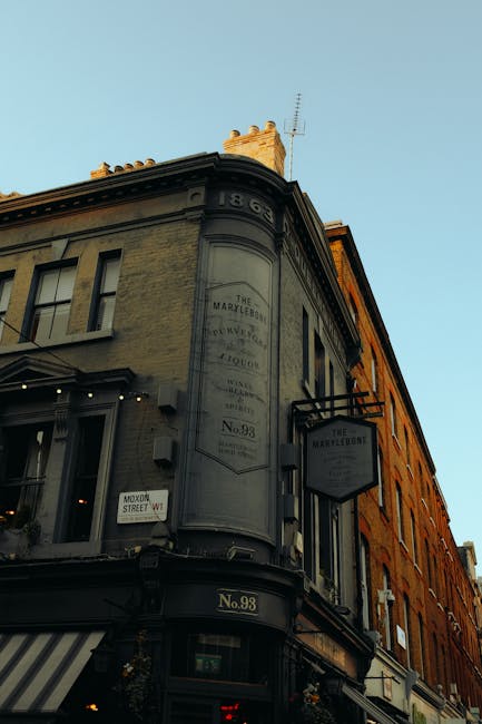 Photograph of a historic multi-storey building on a corner street in Baker Street, Marylebone, with a faded sign indicating the building's name and address, No. 98. The exterior features aged, light-colored stonework with large windows and decorative trim, and a black metal sign hanging from the side of the building displaying 'The Marylebone.' The scene is captured during daylight with a clear blue sky, and nearby shops, including one with a striped awning, are visible at street level. In the foreground, a moving van or removal vehicle, associated with [COMPANY_NAME], is partially visible, indicating a home relocation or furniture transport process. The environment suggests an active commercial area with some interior packing materials and furniture possibly being prepared for moving, aligning with the context of professional removals services in Marylebone, London.