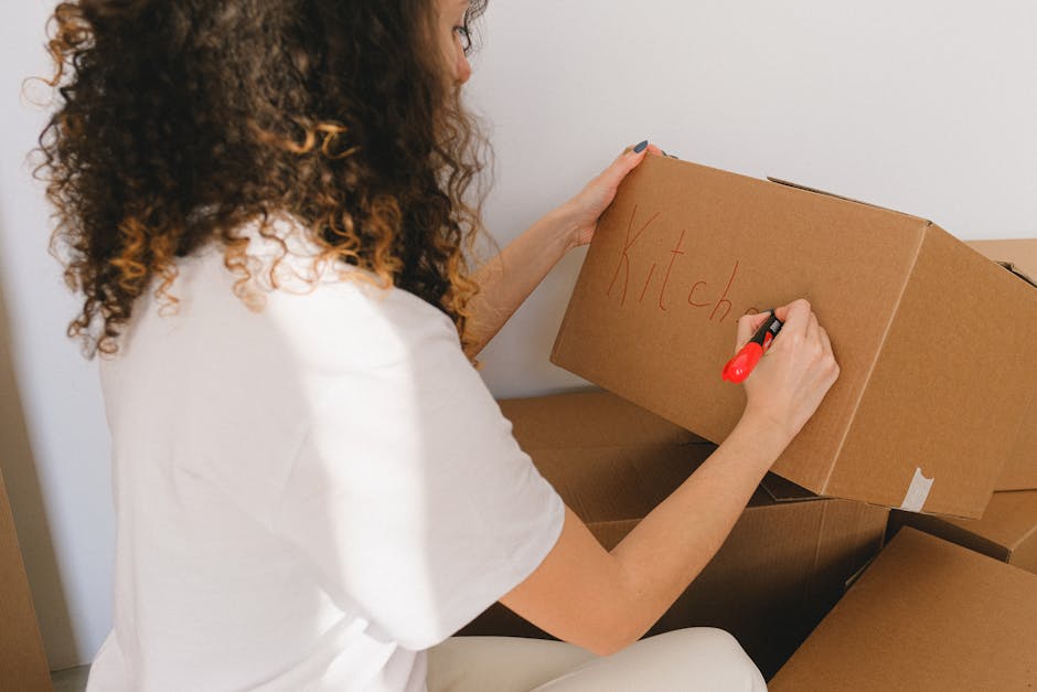 A man and a woman standing inside an entryway near a doorway in an indoor property, each holding a cardboard box used for packing and moving; the man, wearing a maroon t-shirt and orange pants, holds a large box with black tape and shipping labels in front of him, while the woman, dressed in a plaid shirt and beige trousers, holds a smaller box with a red 'Fragile' label. Both are positioned in front of a dark wooden door with the number '200' visible on the wall beside it, and the lighting appears to be natural or from overhead fixtures. The scene depicts the process of home relocation, with the individuals ready for furniture transport or loading as part of a professional removals service by Removal Company Marylebone, supporting efficient packing and moving logistics.
